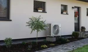 Unidad exterior de aerotermia instalada en casa ya construida. Jardín cuidado con plantas y camino de baldosas.