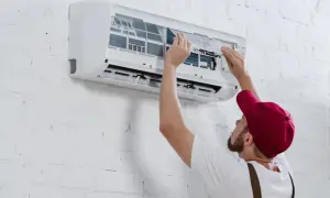 Hombre con gorra roja instala aire acondicionado uno mismo, ajustando filtros en unidad de pared blanca.