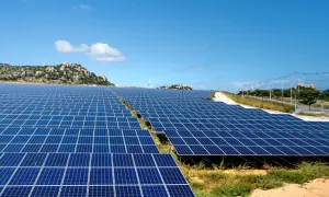 Extenso parque solar con paneles azules bajo un cielo despejado. En el fondo, colinas rocosas y una turbina eólica.