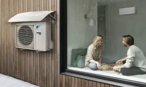 Unidad exterior de cop aerotermia Daikin instalada en fachada de madera, con nieve en el toldo. Dentro, pareja juega en un ambiente cálido.