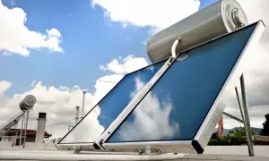 Sistema para calentar agua con placas solares en un tejado, con el cielo azul y nubes reflejándose en los paneles.