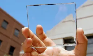 Mano sostiene un panel de placas solares transparentes contra un cielo azul y edificios.