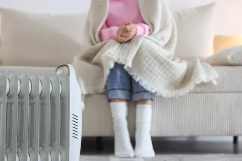 Persona abrigada con manta y calcetines sentada en sofá, junto a un radiador eléctrico. ¿Qué sistema de calefacción elegir para estar cómodo?