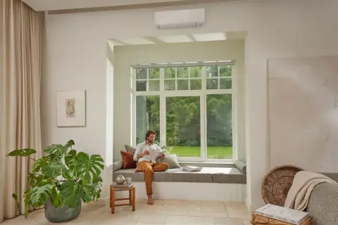 Hombre leyendo en un banco junto a una ventana, con un aire acondicionado instalado arriba. Pregúntate: cuanto se tarda en instalar un aire acondicionado.