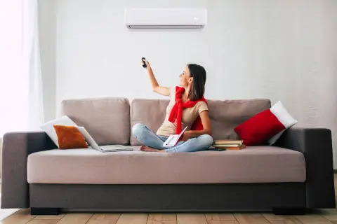 Mujer sentada en sofá, usando control remoto para ajustar uno de los tipos de aire acondicionado.