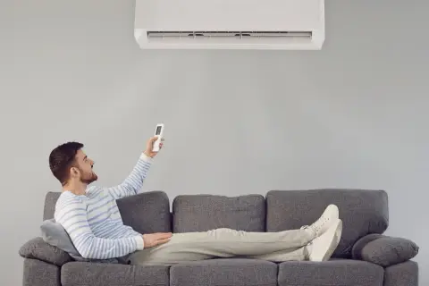Hombre relajado en sofá, usando control remoto para que el aire acondicionado renueve el aire y refresque la estancia.