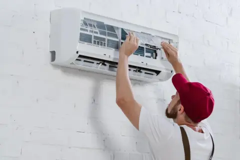 Hombre con gorra roja instala aire acondicionado uno mismo, ajustando filtros en unidad de pared blanca.