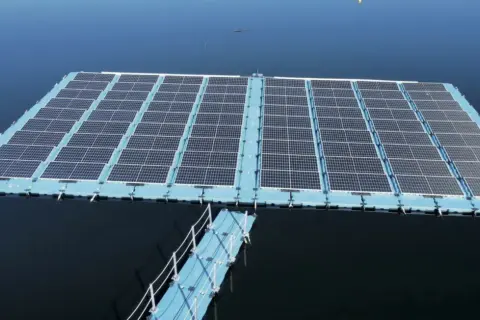 Plataforma de energía fotovoltaica flotante sobre un cuerpo de agua azul, con un pasarela azul conectada.