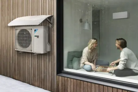 Unidad exterior de cop aerotermia Daikin instalada en fachada de madera, con nieve en el toldo. Dentro, pareja juega en un ambiente cálido.