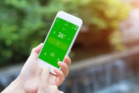 Mano usando un móvil para controlar el aire acondicionado, mostrando la temperatura de 26 grados en la pantalla verde.