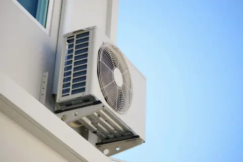 Un aire acondicionado en balcon, montado en la pared blanca de un edificio, con el cielo azul de fondo.