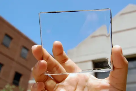 Mano sostiene un panel de placas solares transparentes contra un cielo azul y edificios.
