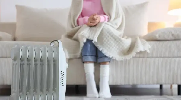 Persona abrigada con manta y calcetines sentada en sofá, junto a un radiador eléctrico. ¿Qué sistema de calefacción elegir para estar cómodo?