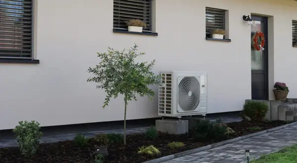 Unidad exterior de aerotermia instalada en casa ya construida. Jardín cuidado con plantas y camino de baldosas.