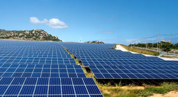 Extenso parque solar con paneles azules bajo un cielo despejado. En el fondo, colinas rocosas y una turbina eólica.