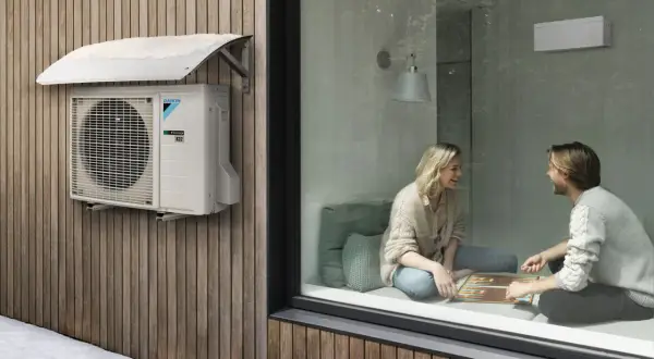 Unidad exterior de cop aerotermia Daikin instalada en fachada de madera, con nieve en el toldo. Dentro, pareja juega en un ambiente cálido.