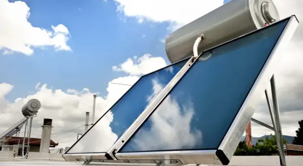 Sistema para calentar agua con placas solares en un tejado, con el cielo azul y nubes reflejándose en los paneles.