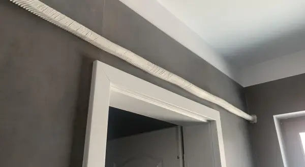Ideas para tapar tubos aire acondicionado: un tubo blanco texturizado recorre la pared gris sobre un marco de puerta blanco.