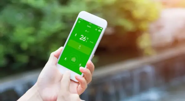 Mano usando un móvil para controlar el aire acondicionado, mostrando la temperatura de 26 grados en la pantalla verde.