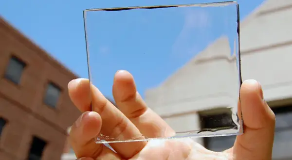Mano sostiene un panel de placas solares transparentes contra un cielo azul y edificios.