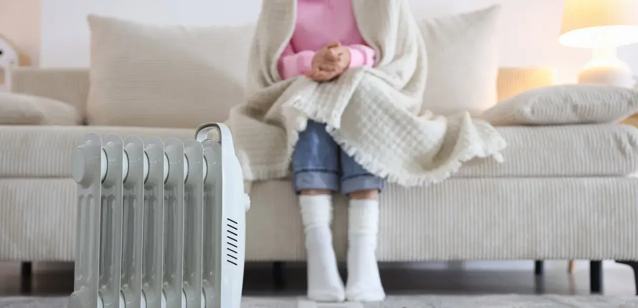 Persona abrigada con manta y calcetines sentada en sofá, junto a un radiador eléctrico. ¿Qué sistema de calefacción elegir para estar cómodo?