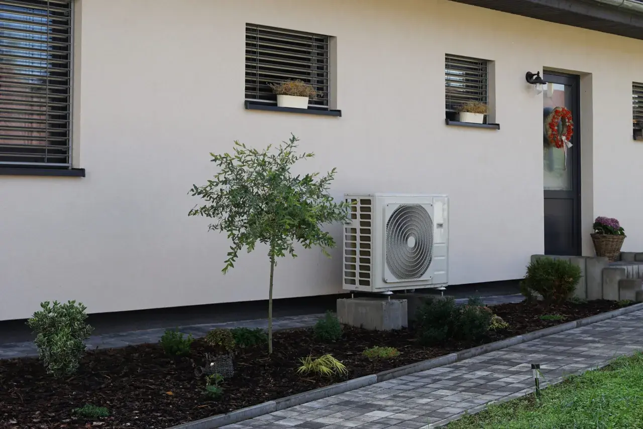 Unidad exterior de aerotermia instalada en casa ya construida. Jardín cuidado con plantas y camino de baldosas.