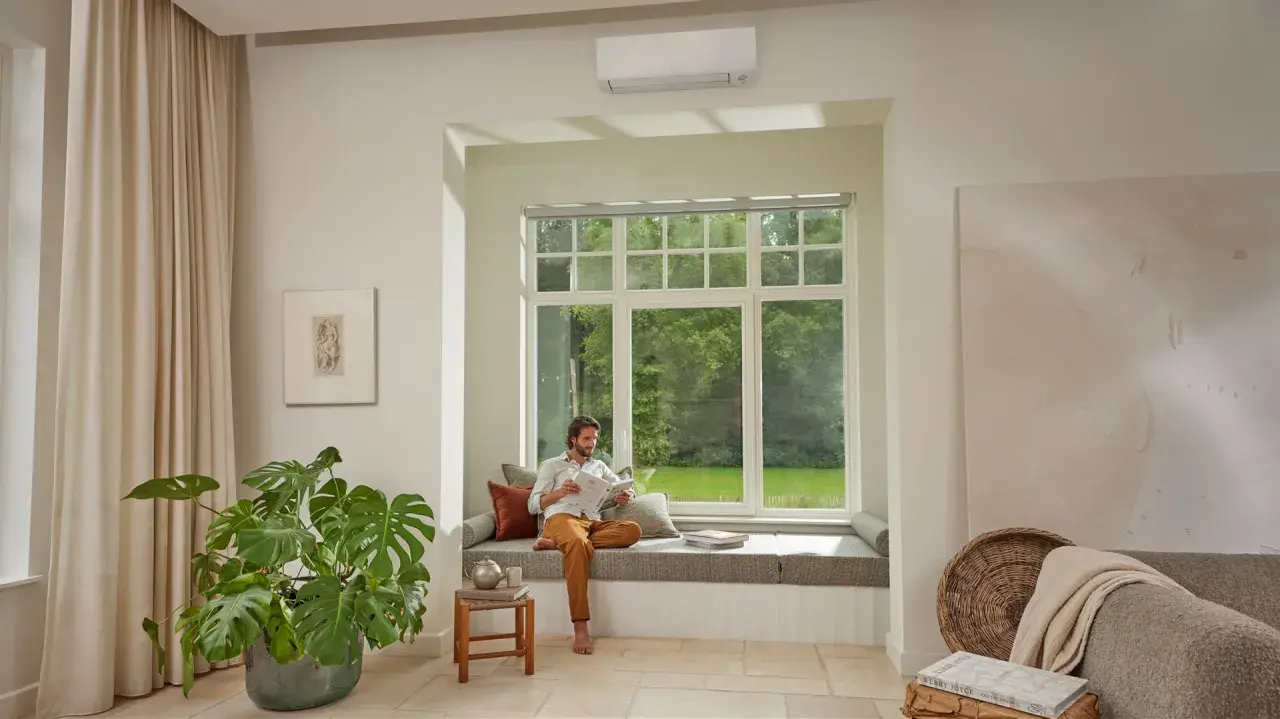 Hombre leyendo en un banco junto a una ventana, con un aire acondicionado instalado arriba. Pregúntate: cuanto se tarda en instalar un aire acondicionado.
