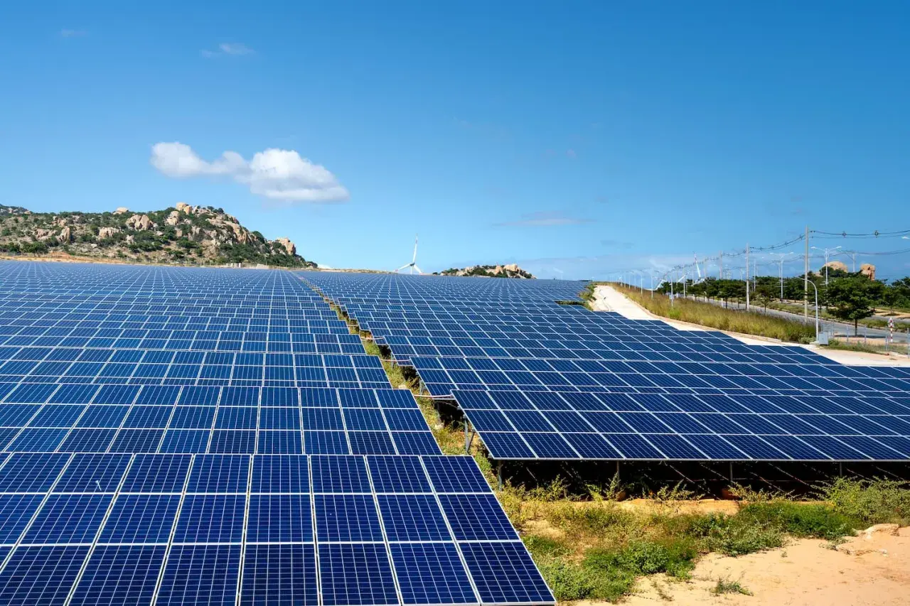 Extenso parque solar con paneles azules bajo un cielo despejado. En el fondo, colinas rocosas y una turbina eólica.
