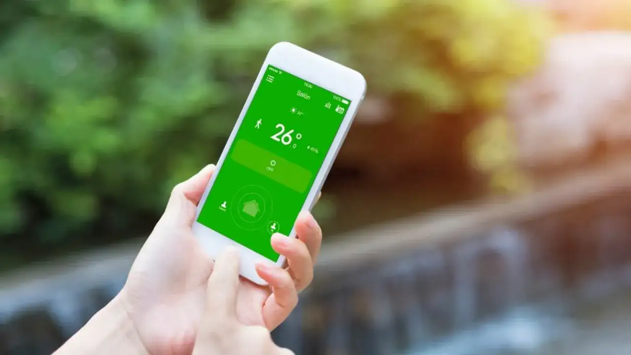 Mano usando un móvil para controlar el aire acondicionado, mostrando la temperatura de 26 grados en la pantalla verde.