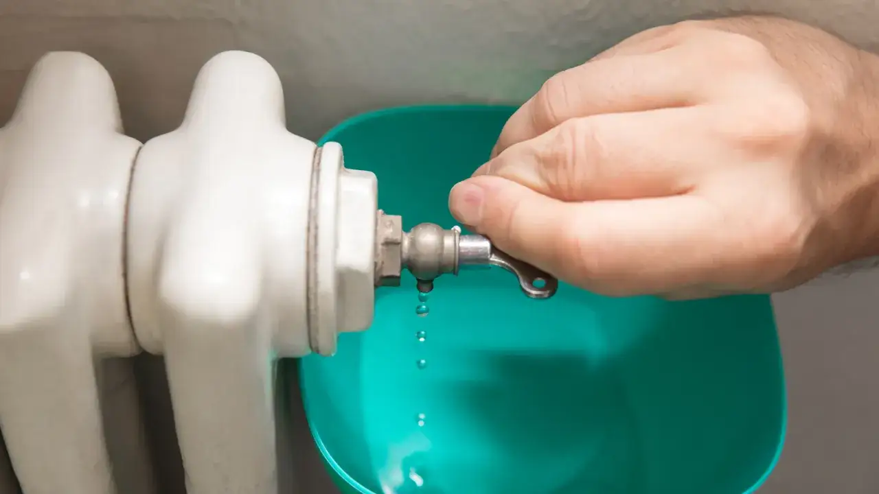 Mano girando la llave para purgar radiadores, goteando agua en un recipiente verde.