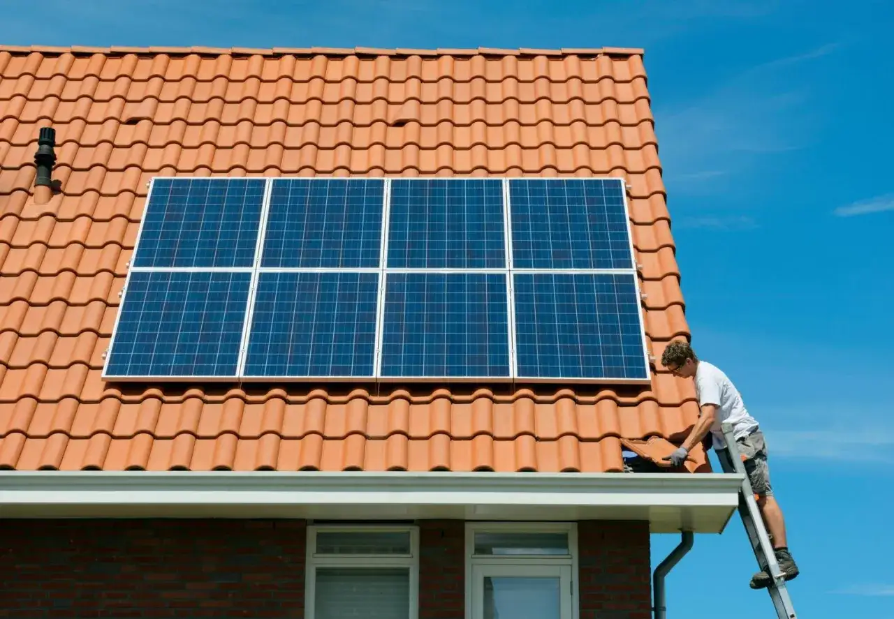 Un técnico en una escalera realiza la instalación de placas solares en un tejado de tejas naranjas bajo un cielo azul.
