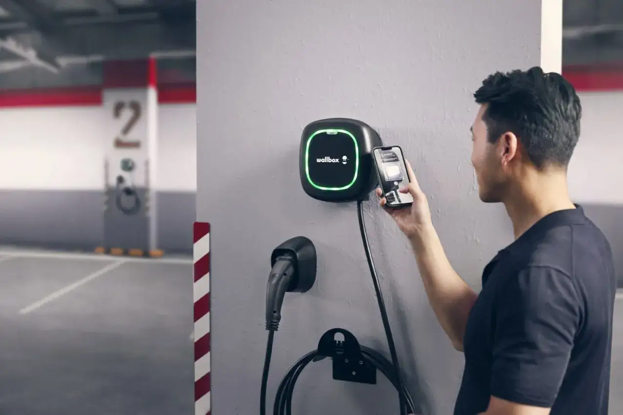 Hombre usa móvil para instalar punto de recarga en garaje comunitario. El cargador Wallbox se ilumina en verde.