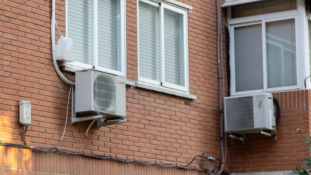 Dos unidades exteriores de instalación aire acondicionado en fachada de ladrillo, junto a ventanas con persianas.