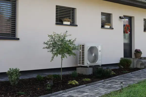 Unidad exterior de aerotermia instalada en casa ya construida. Jardín cuidado con plantas y camino de baldosas.