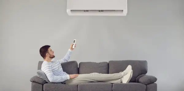 Hombre relajado en sofá, usando control remoto para que el aire acondicionado renueve el aire y refresque la estancia.