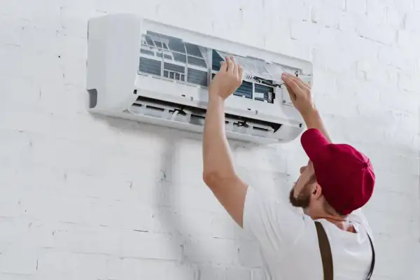 Hombre con gorra roja instala aire acondicionado uno mismo, ajustando filtros en unidad de pared blanca.