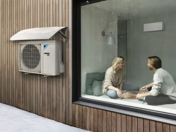 Unidad exterior de cop aerotermia Daikin instalada en fachada de madera, con nieve en el toldo. Dentro, pareja juega en un ambiente cálido.