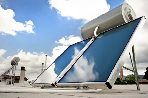 Sistema para calentar agua con placas solares en un tejado, con el cielo azul y nubes reflejándose en los paneles.