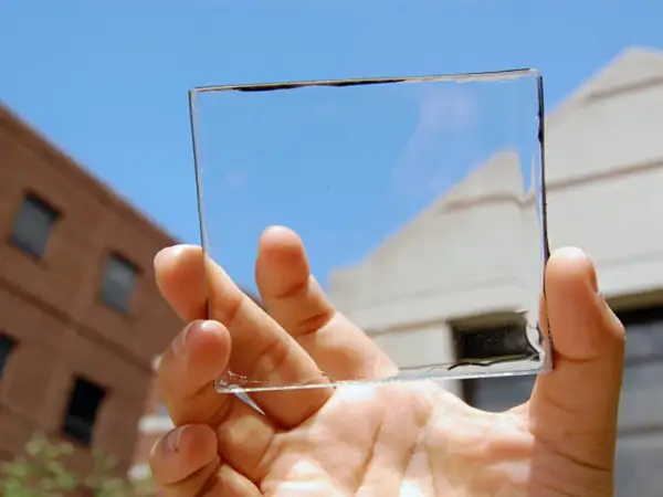 Mano sostiene un panel de placas solares transparentes contra un cielo azul y edificios.